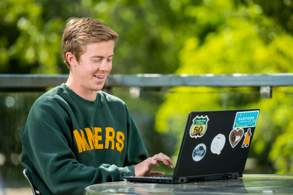 A student uses his laptop at a table outside, while wearing a MINERS sweatshirt.